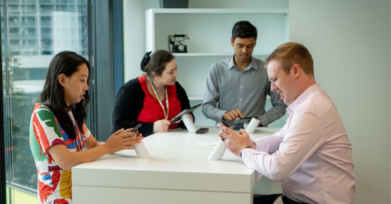 Four office employees sitting at desk using technical devices